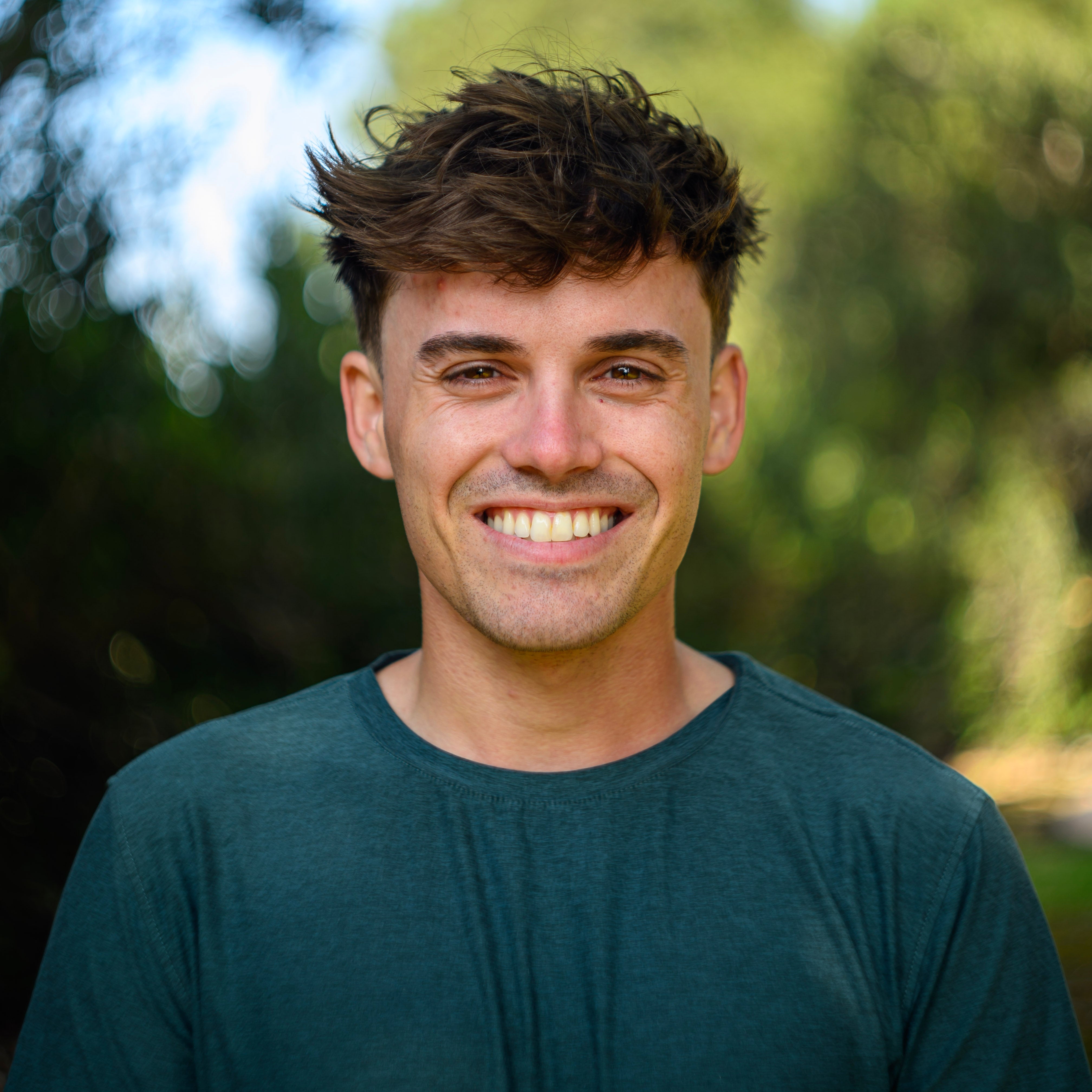 A smiling man wearing a green shirt, standing outdoors with a blurred natural background. Captured with a high-resolution camera, this portrait showcases sharp image quality, natural lighting, and a soft bokeh effect for professional photography applications.